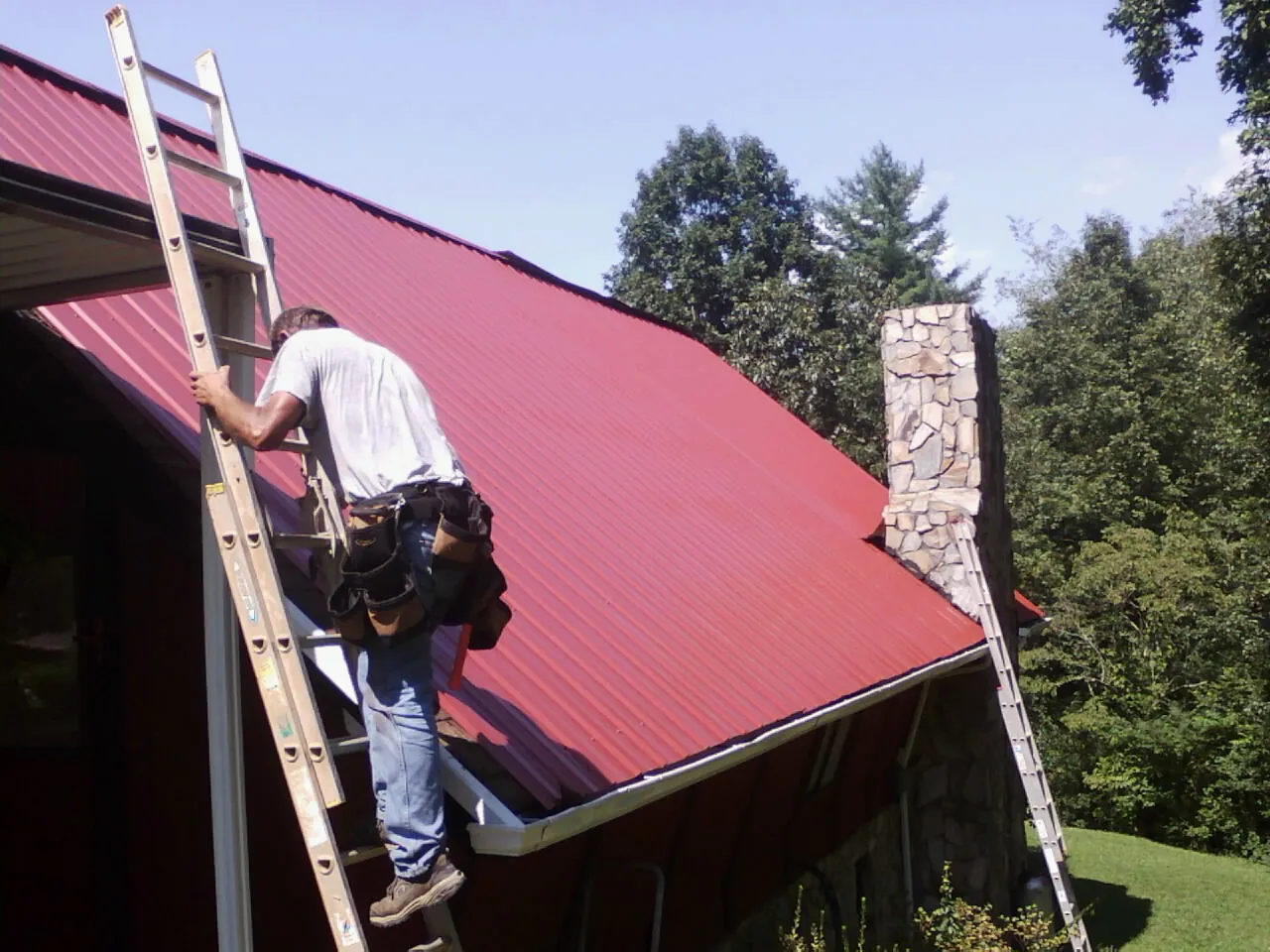 Roofer on a ladder beside a red metal roof and stone chimney — Soffit & Fascia Repair by Roofing Brothers in Hanford